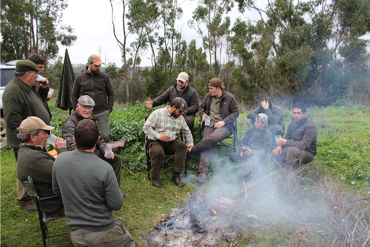 Comensalismo (Paterna del Campo, Huelva)
La comida que culmina la monter&iacute;a, bien en la sede de las sociedades y pe&ntilde;as, bien en campo, como atestigua esta imagen, supone el momento de disfrute de los lazos sociales, ampliando la comunidad montera a familiares y amigos de los cazadores. El modelo que atestigua esta imagen es el de lo que se conoce como &ldquo;monter&iacute;a social&rdquo;, que se remata con un almuerzo en el campo, dispuesto con las aportaciones de cada grupo familiar. Embutidos, tortillas y carne son alimentos imprescindibles en estos almuerzos, as&iacute; como guisos monteros, realizados a base de carne de caza. Es una peculiaridad de esta zona la caldereta de jabal&iacute;, incluyendo el &ldquo;corato&rdquo;, nombre que recibe el duro pellejo de este animal que queda muy gelatinoso en el guiso. Los aficionados de esta sociedad celebran la caldereta con corato por encima de cualquier otra vianda.
Conforme pasan las horas, las conversaciones sobre la jornada de caza van dando paso a la recuperaci&oacute;n de recuerdos, a las bromas y chanzas. Entretanto, van volviendo los perros de las rehalas y, llegado un punto de la tarde, antes de la oscurecida, la mesa donde se han sentado a comer los perreros inicia el momento del cante. Surgen fandangos de distintas voces que se suceden, cuyas letras recrean el mundo de la caza, haciendo emerger un momento.